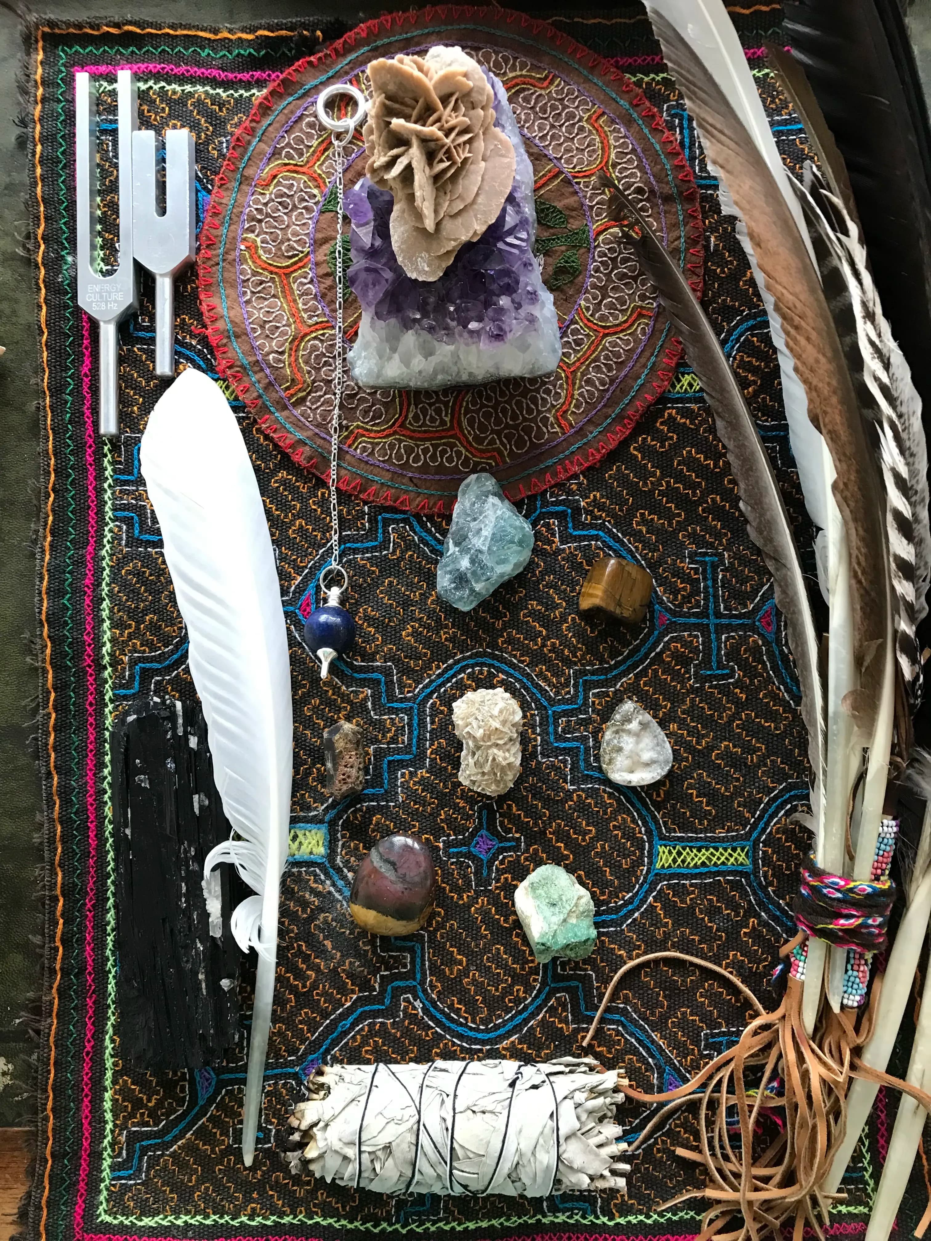 Crystals, feathers, tuning forks and sage laid out on an embroidered cloth
