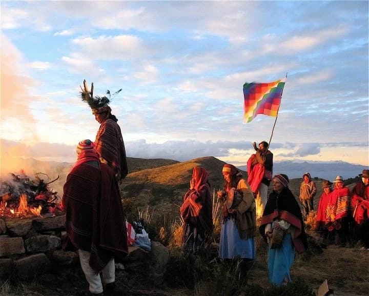 A fire ceremony at dusk in the Andes, people in ponchos around the flames under a wiphala flag