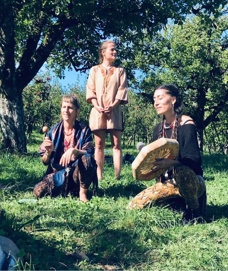 Three women in an orchard, one playing a frame drum, preparing a ceremony